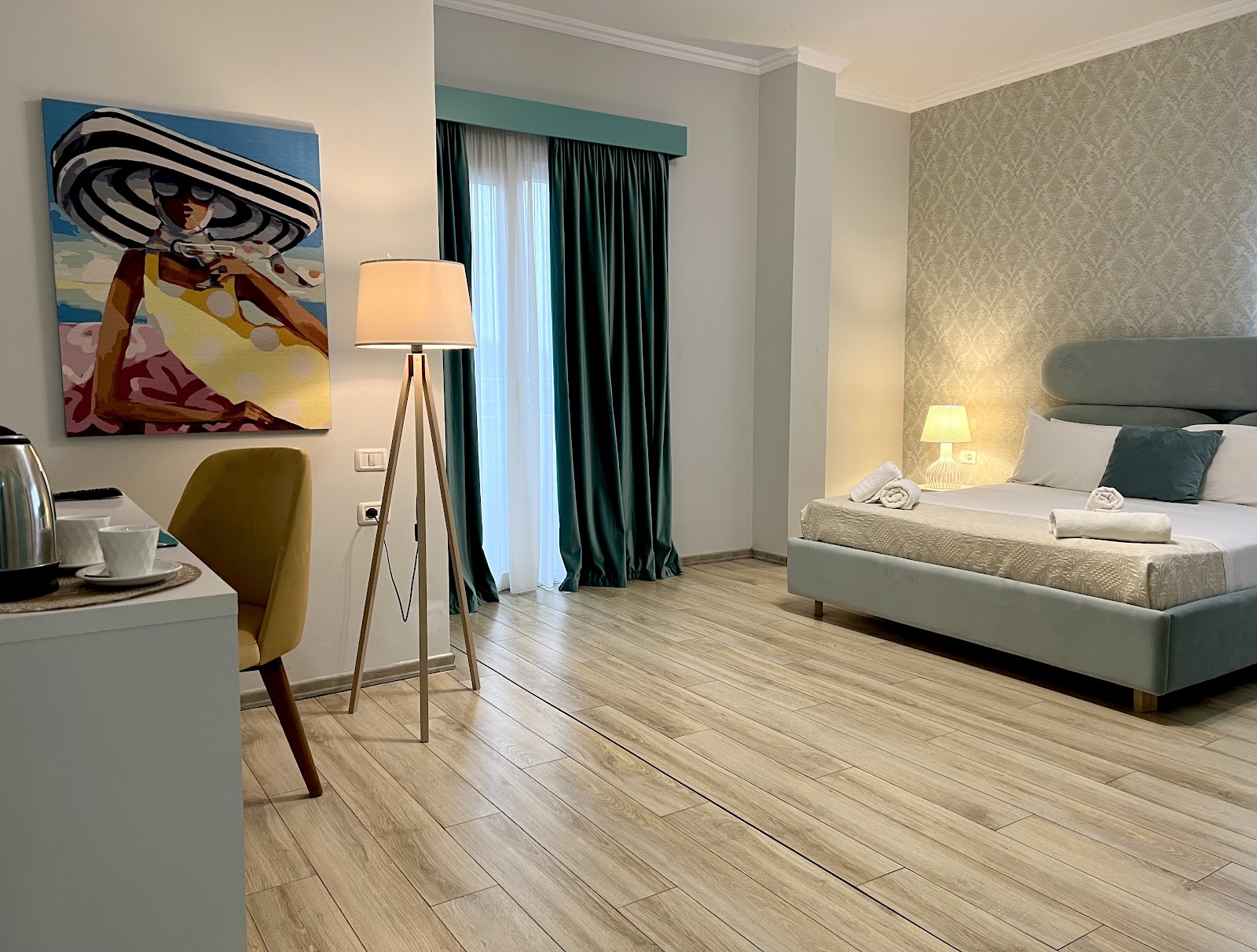 Resort guest room with queen bed, green headboard, patterned wallpaper, wooden flooring, and beach-themed artwork above a work desk.