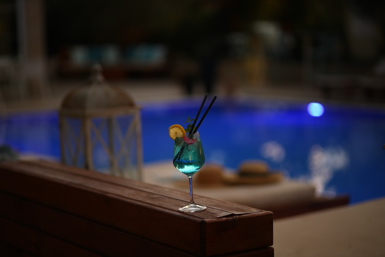 Turquoise cocktail with lemon slice and black straws sits on wooden railing overlooking MarBella Beach Hotel's illuminated pool at dusk.