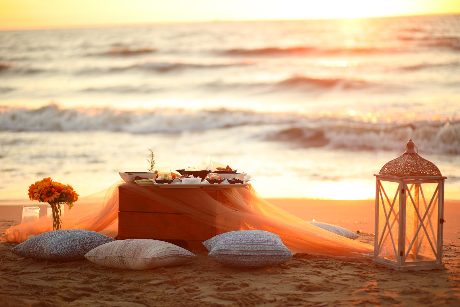 Sunset beach picnic setup with pillows, food platters, orange fabric draping, lantern, and flowers at MarBella Beach Hotel.