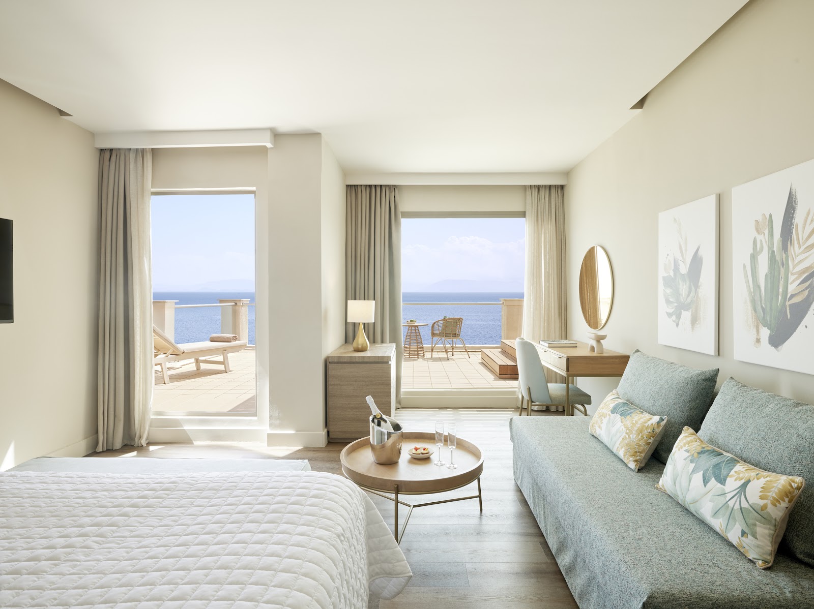 Bright resort bedroom with white bed, wooden desk, and round mirror, opening to ocean-view terrace with lounge chairs.