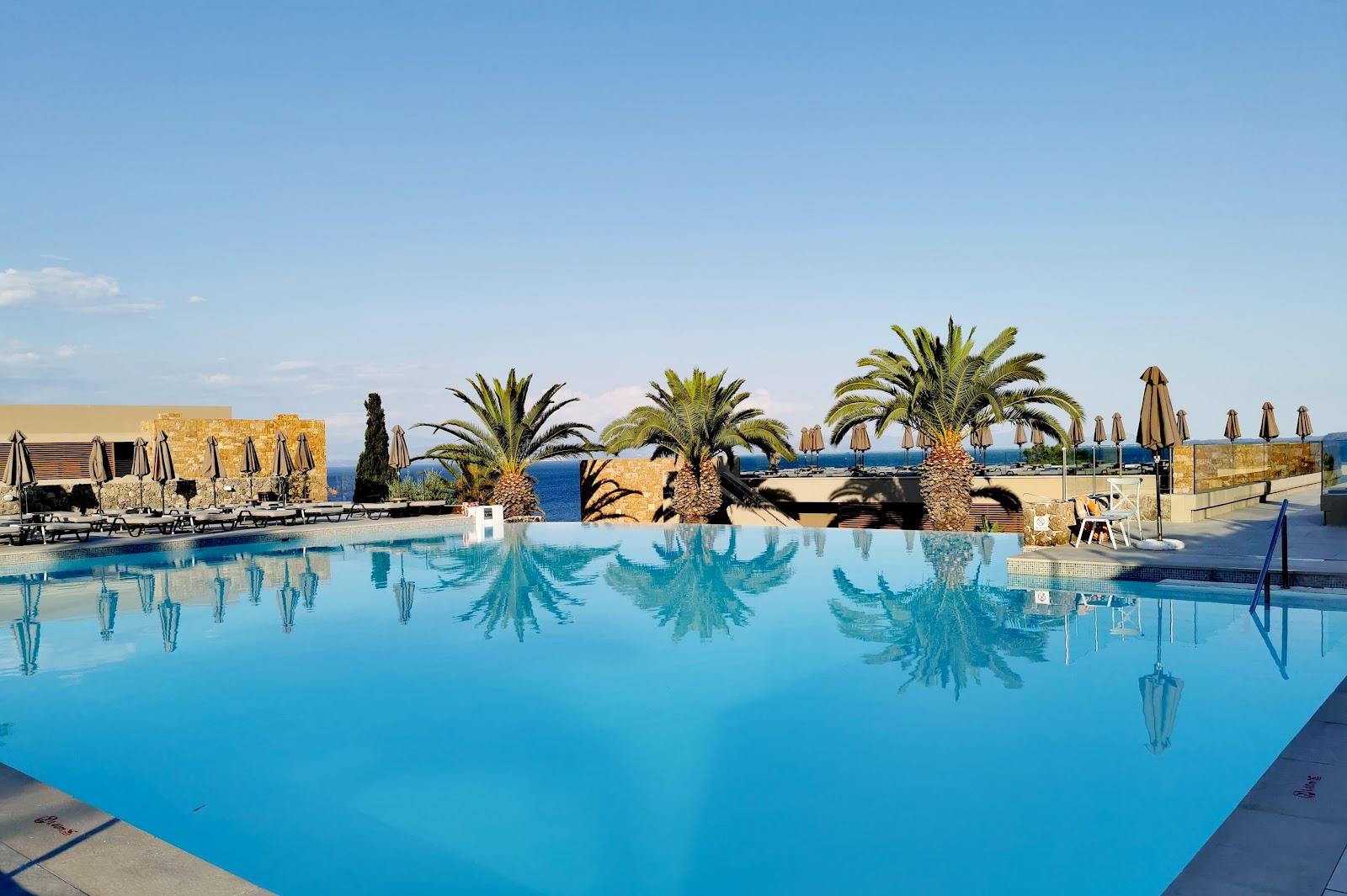 Large infinity pool with loungers and umbrellas at MarBella, stone buildings and palm trees visible at dusk with cloudy sky.