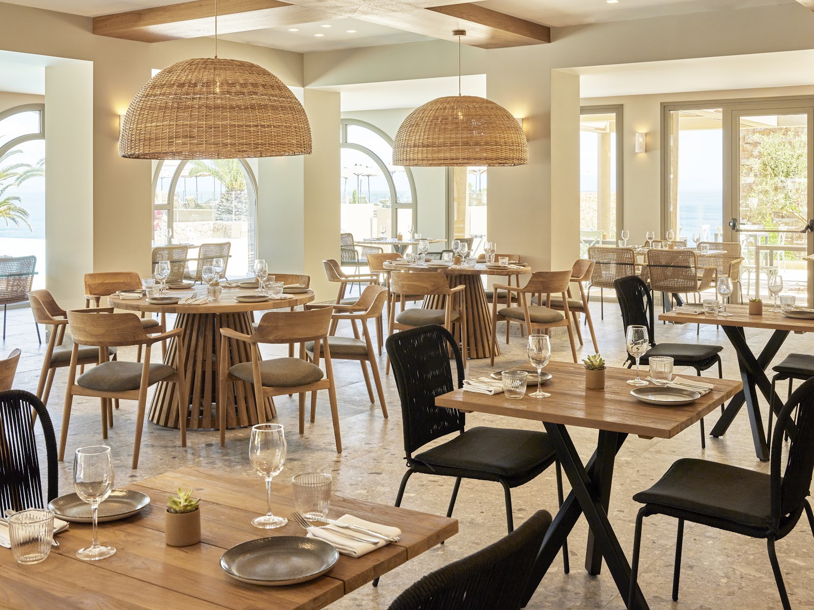 Resort dining room with woven pendant lights, wooden tables, mixed seating, and arched windows overlooking the sea at MarBella.