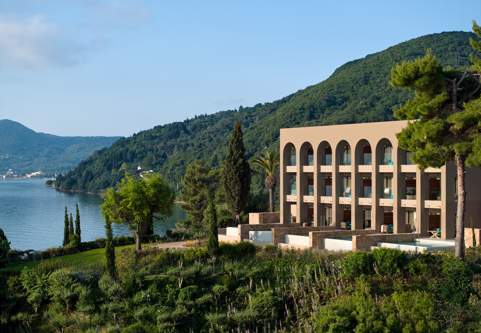 Modern beige building with arched windows overlooks a lake surrounded by forested hills at MarBella resort.