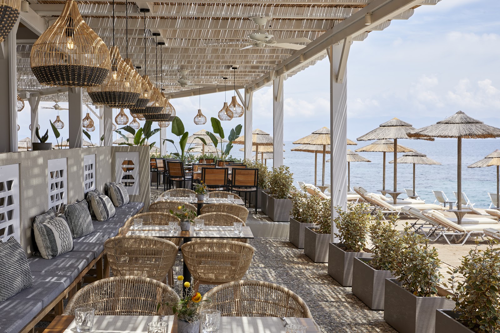 Beachfront dining terrace at MarBella with woven pendant lights, white pergola, wicker chairs, and thatched umbrellas overlooking the sea.