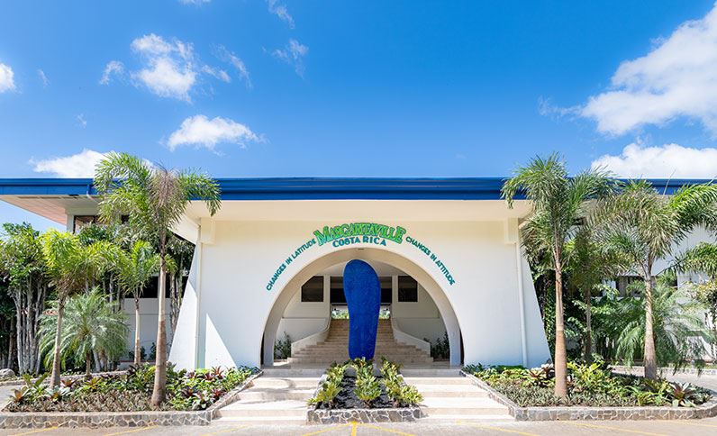 White building entrance with large blue arched doorway, palm trees, and tropical landscaping at Margaritaville Beach Resort Playa Flamingo under blue sky.