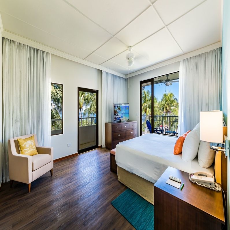 Resort bedroom with king bed, armchair, dark wood floors, and balcony overlooking palm trees at Margaritaville Beach Resort Playa Flamingo.