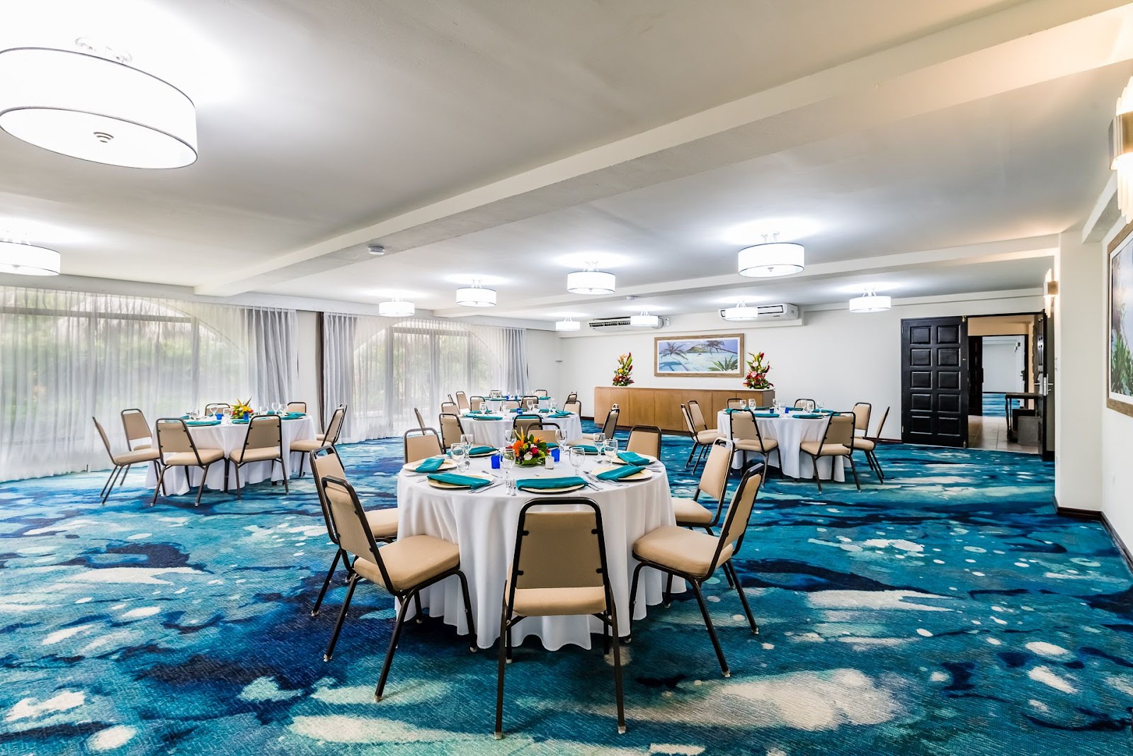 Ballroom with round tables set with white linens and turquoise place settings, tan chairs, blue patterned carpet, and floor-to-ceiling windows.