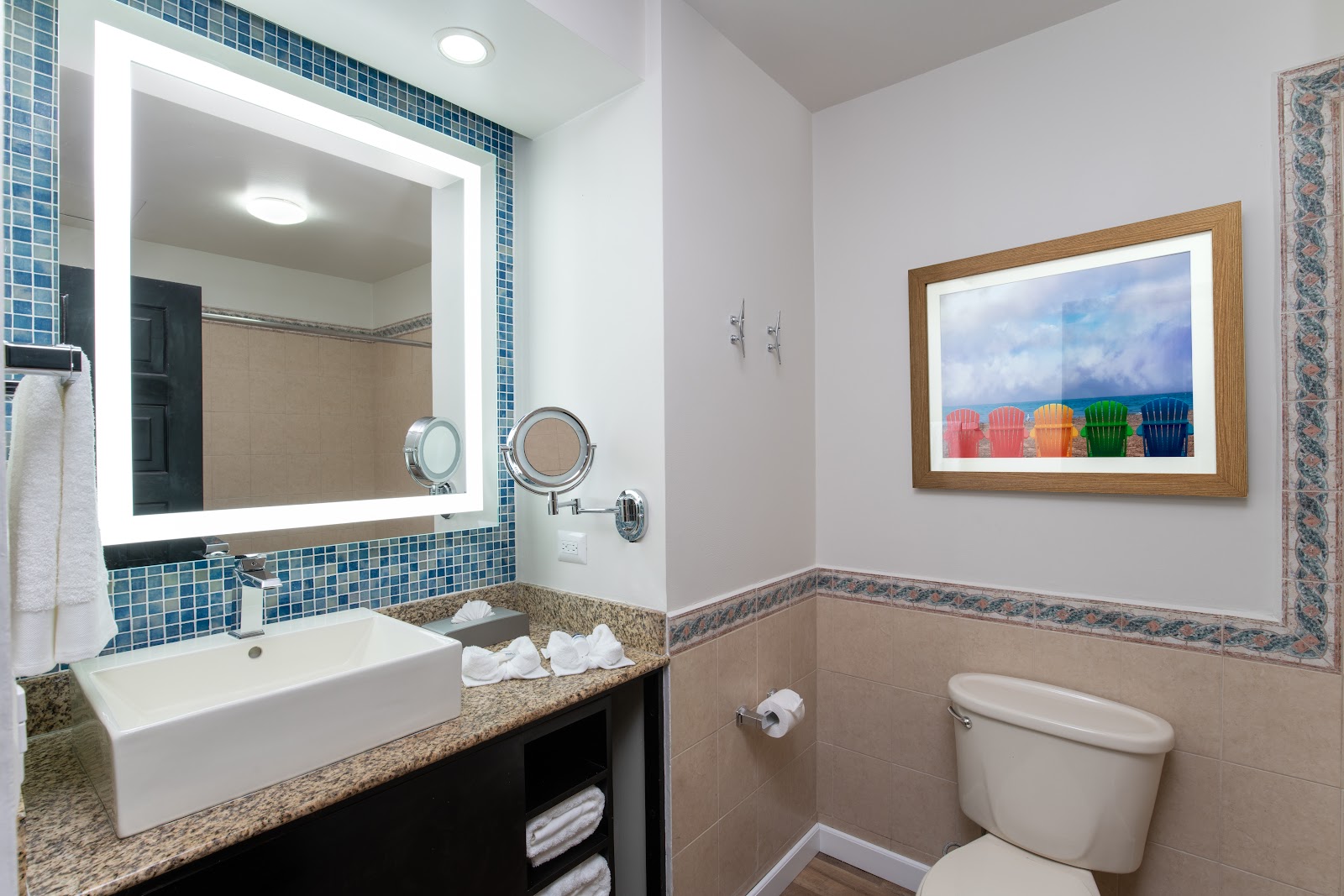 Bathroom with blue tile backsplash, lighted mirror, granite countertop, and artwork of colorful beach chairs on the wall.