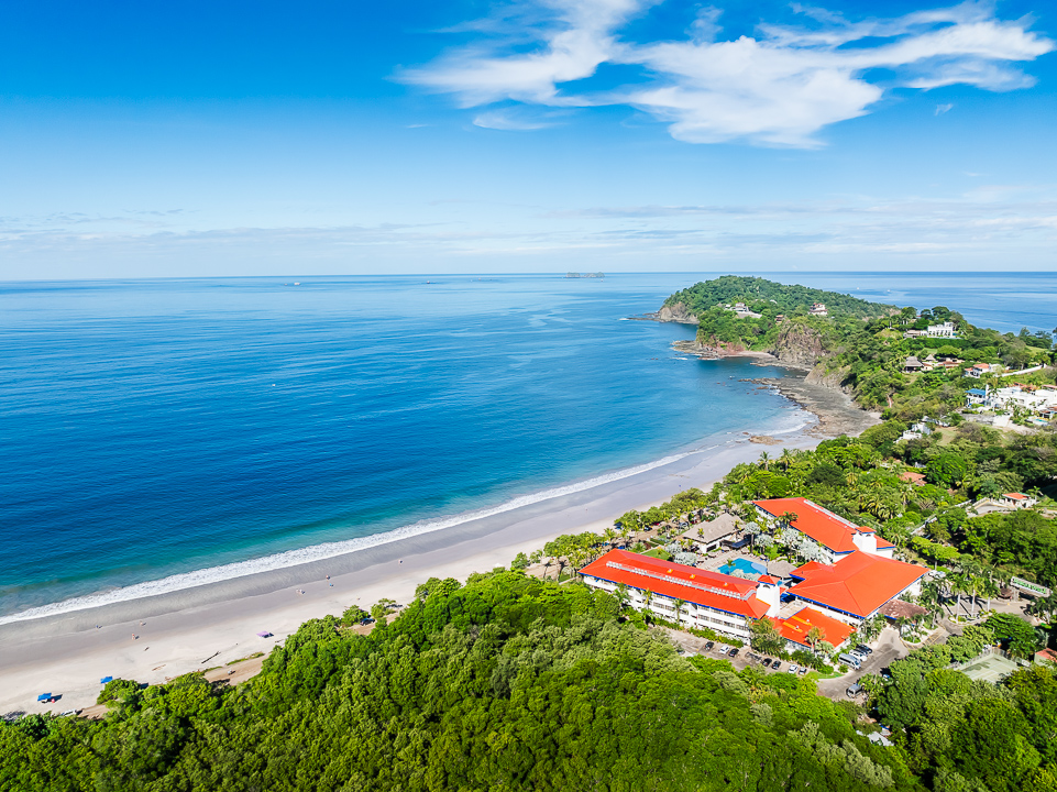 Aerial view of Margaritaville Beach Resort Playa Flamingo with orange roofs nestled between dense green forest and a sandy beach along turquoise ocean waters.