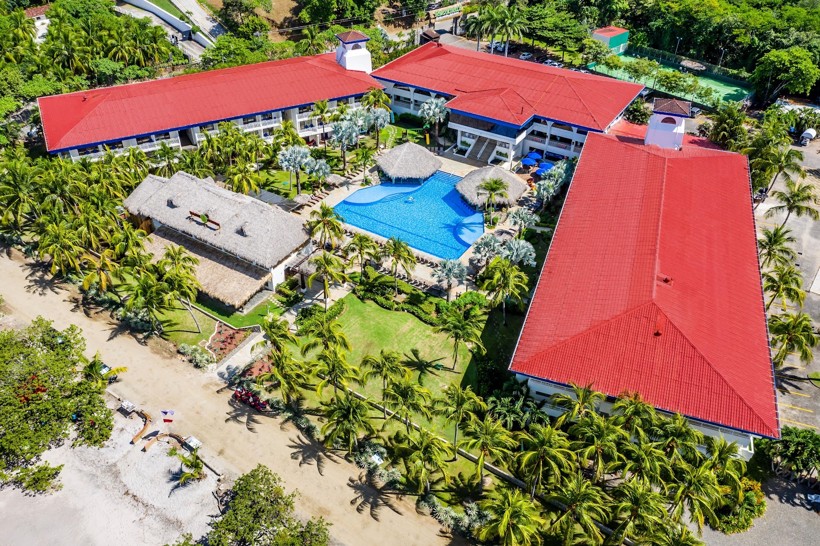 Aerial view of Margaritaville Beach Resort Playa Flamingo with red-roofed buildings, blue pool, thatched-roof palapas, palm trees, and sandy beach.