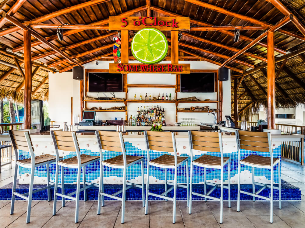 Bar seating at 5 O'Clock Somewhere Bar with wooden beam ceiling, lime slice sign, and blue tiled counter at Margaritaville Beach Resort Playa Flamingo.