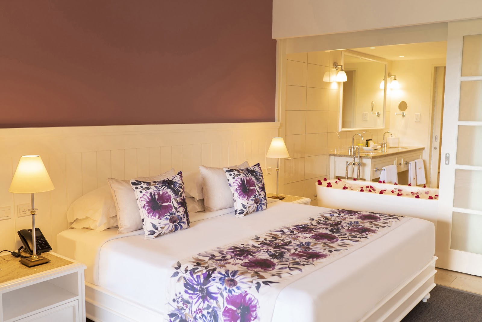 Resort bedroom with king bed featuring purple floral bedding, matching pillows, and rose petals in soaking tub visible through open bathroom door.