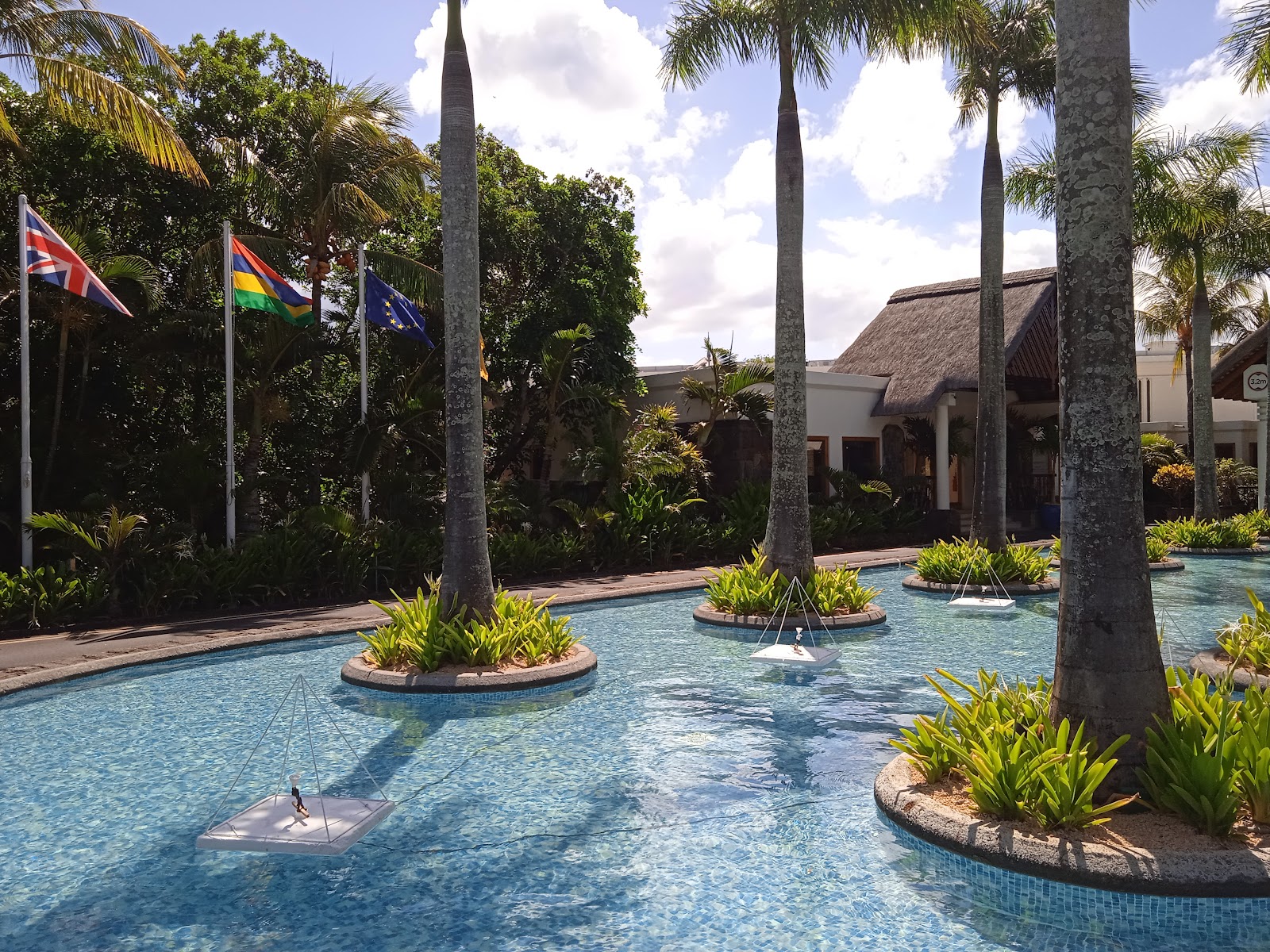 Shallow lagoon-style pool with tall palm trees, tropical plants, and international flags near thatched-roof buildings under blue sky.