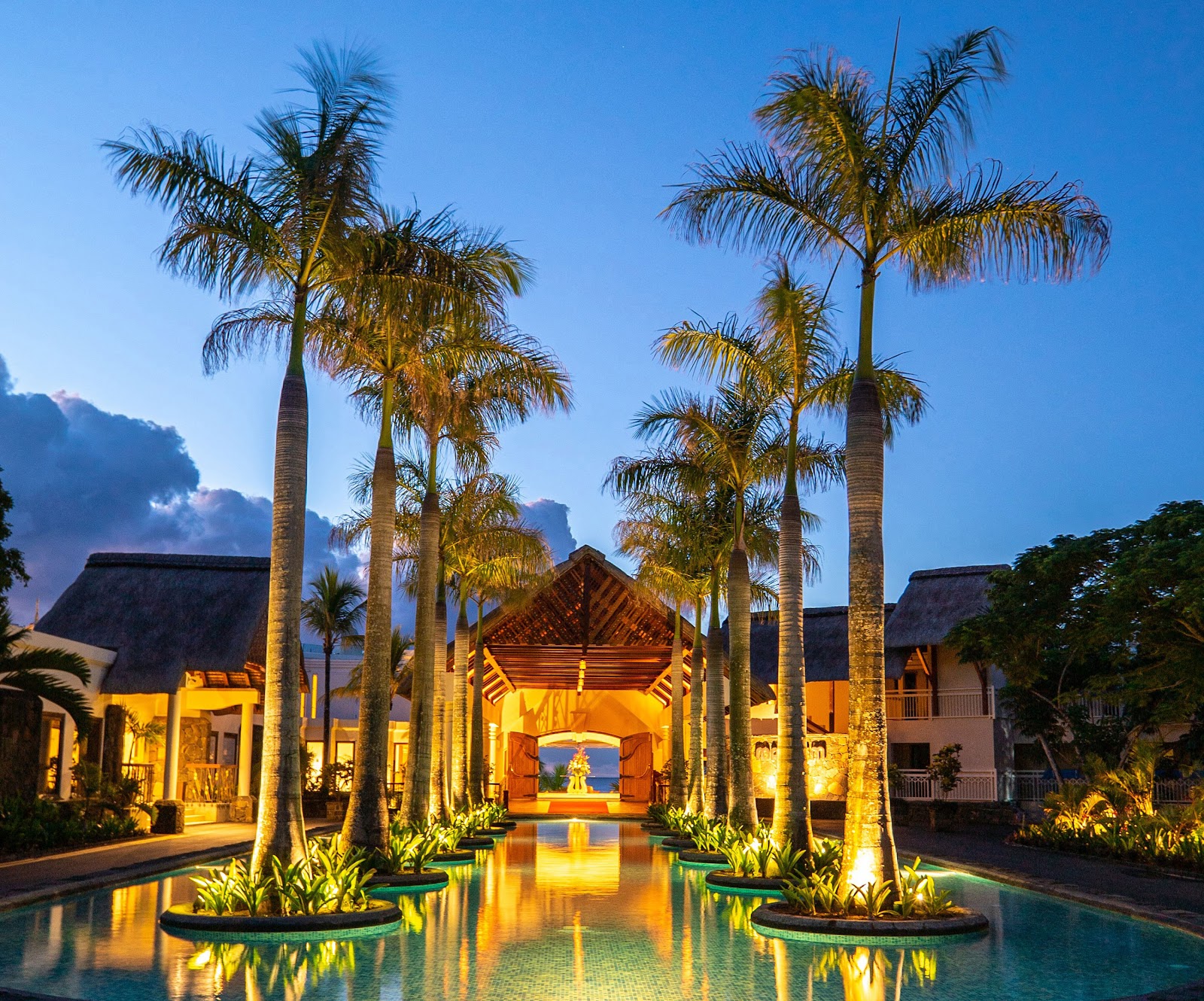 Illuminated resort courtyard at dusk with tall palms flanking a reflecting pool leading to a lit pavilion, ocean visible beyond.