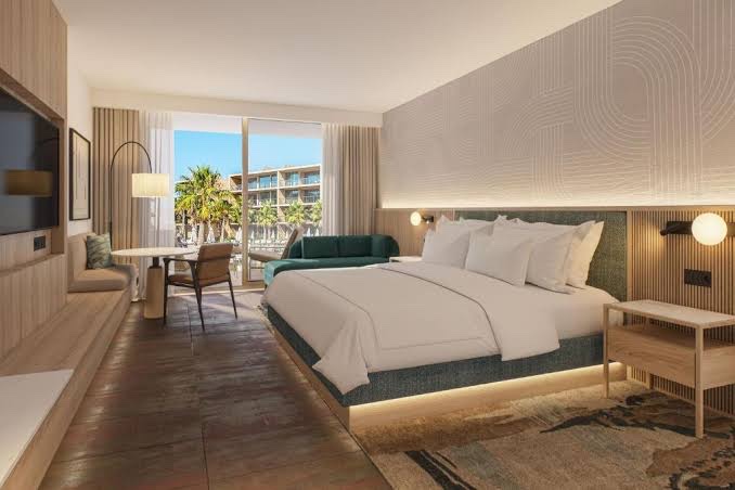 Modern resort bedroom with king bed, desk area, and balcony overlooking palm trees and ocean-view buildings at Marriott Residences Salgados.