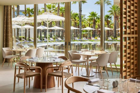 Indoor dining area with round wood tables and beige chairs overlooking a pool, palm trees, and white umbrellas at Marriott Residences Salgados Resort.