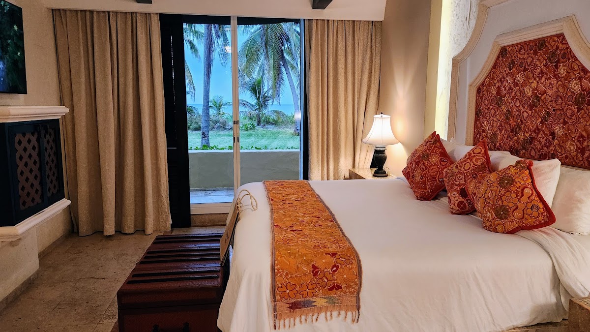 Resort bedroom with white bed topped with red and gold patterned pillows and runner, sliding glass doors opening to ocean and palm trees beyond.