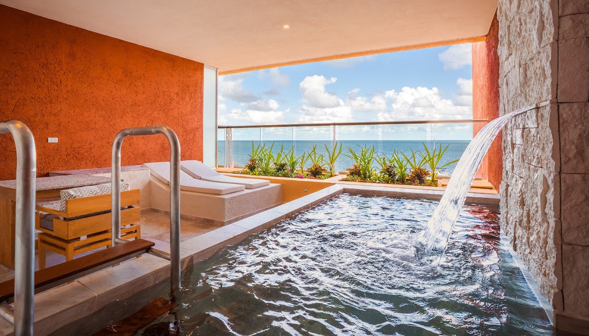 Spa pool with waterfall and loungers overlooking the ocean at Marriott All-Inclusive Riviera Maya under blue sky.