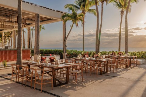 Wooden dining tables and chairs arranged on a beachfront terrace beneath tall palm trees at sunset.