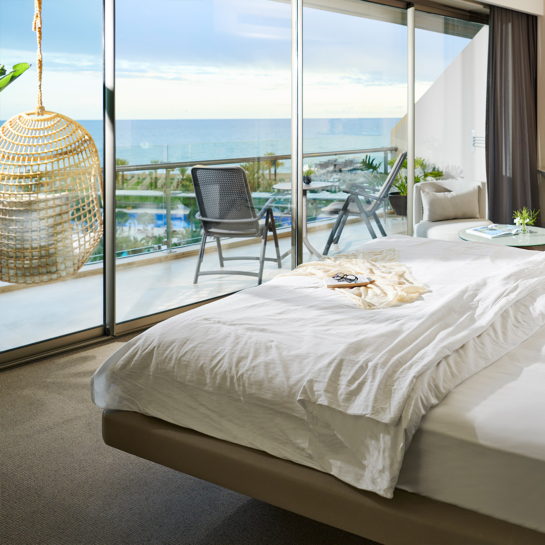 Bedroom with white linens and a woven pendant lamp overlooks a sea-view balcony with lounge chairs and pool at Maxx Royal Belek Golf Resort.