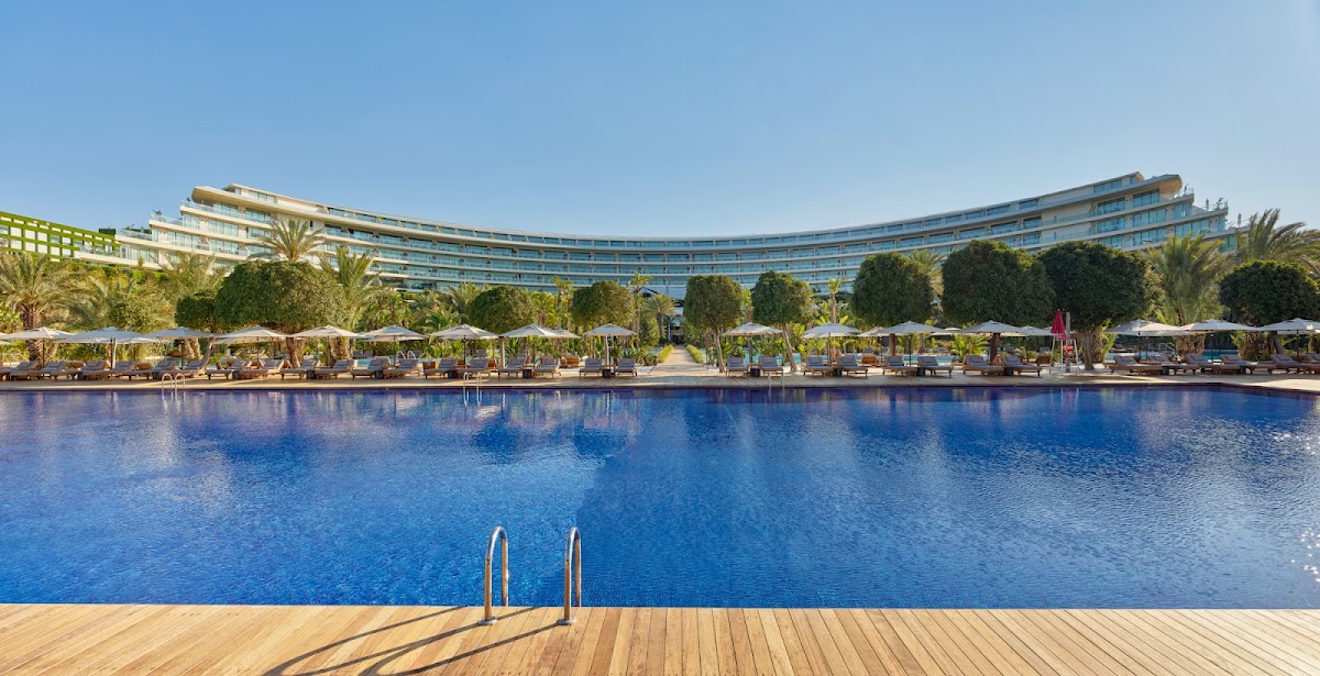 Large curved resort building overlooks a blue pool with wooden deck, lounge chairs, umbrellas, and palm trees lining the water.