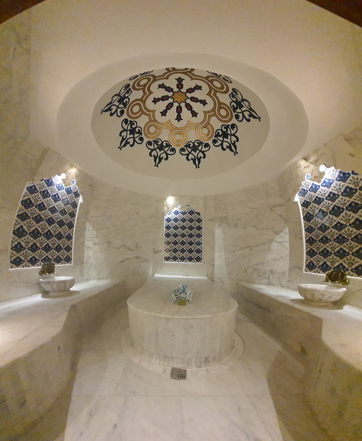 Marble spa interior with domed ceiling featuring intricate geometric medallion, patterned windows, and circular stone basin in center.