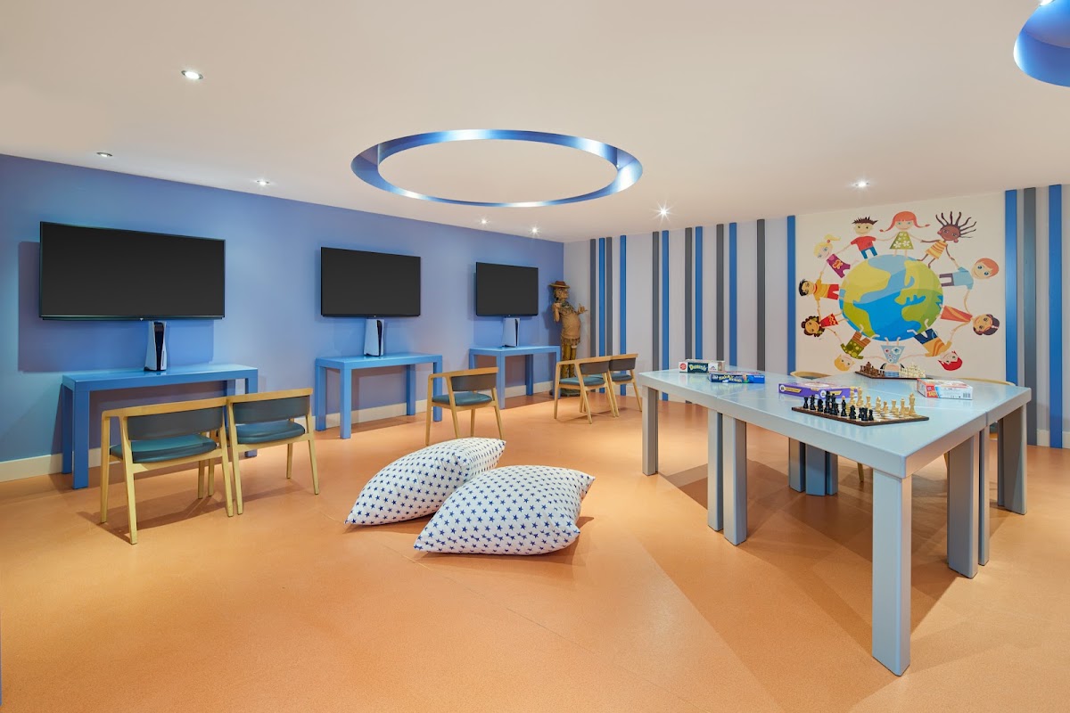 Kids club room with blue walls, activity tables, floor cushions, screens for entertainment, and children's artwork on the wall.