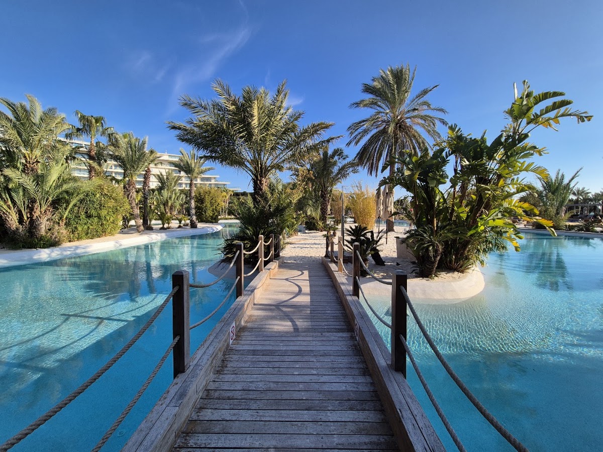 Wooden boardwalk lined with rope railings stretches between turquoise pools surrounded by palm trees at Maxx Royal Belek Golf Resort.