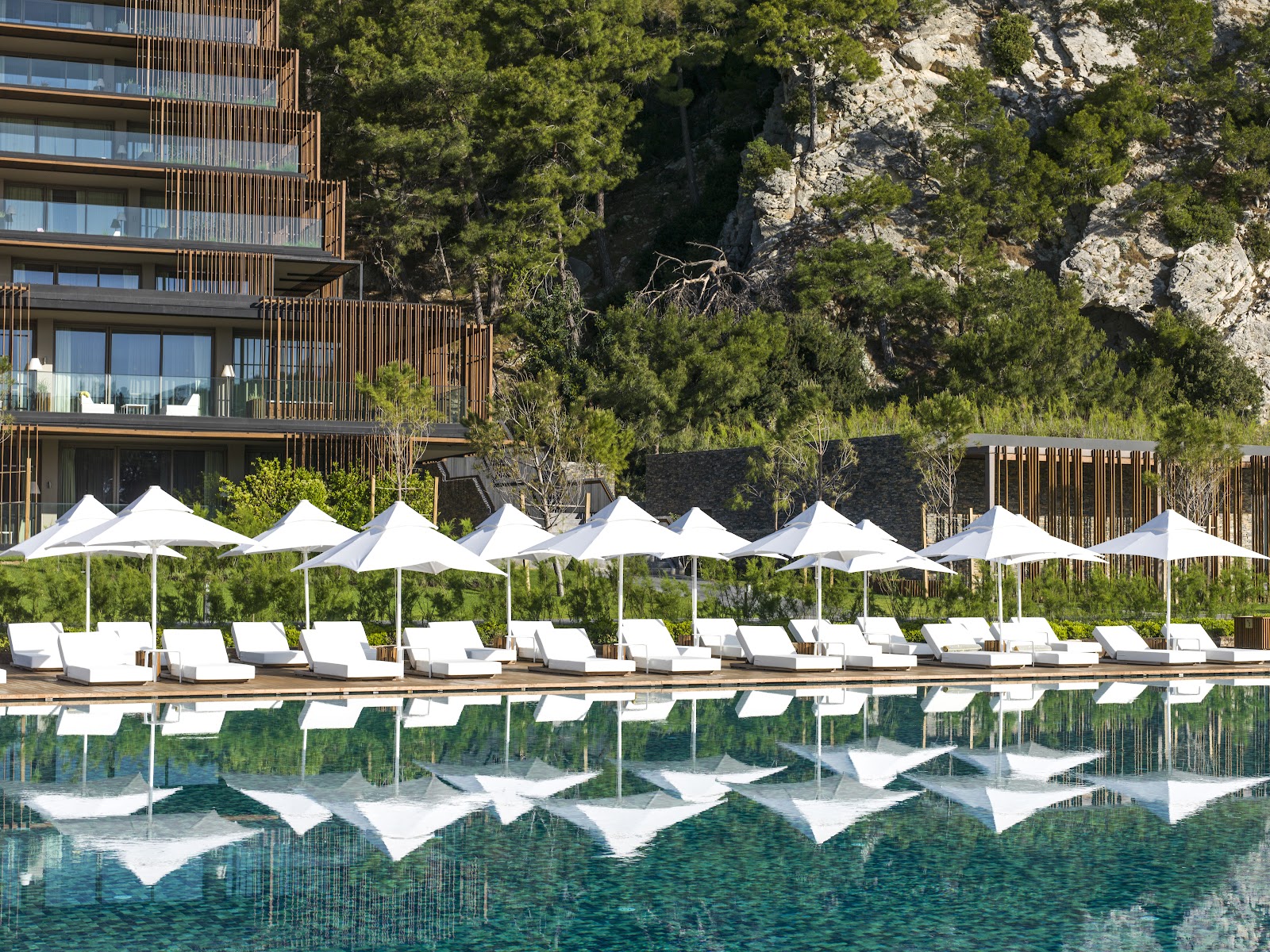 Modern resort building with white sunbeds and canopies lining a pool, surrounded by pine trees and limestone cliffs.