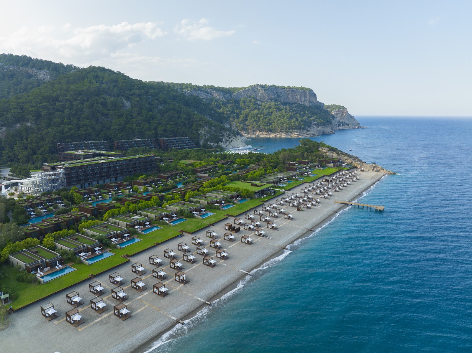 Maxx Royal Kemer Resort's beach with rows of cabanas on sand, green lawns with pools, dark buildings on cliff, and forested hillside overlooking turquoise sea.