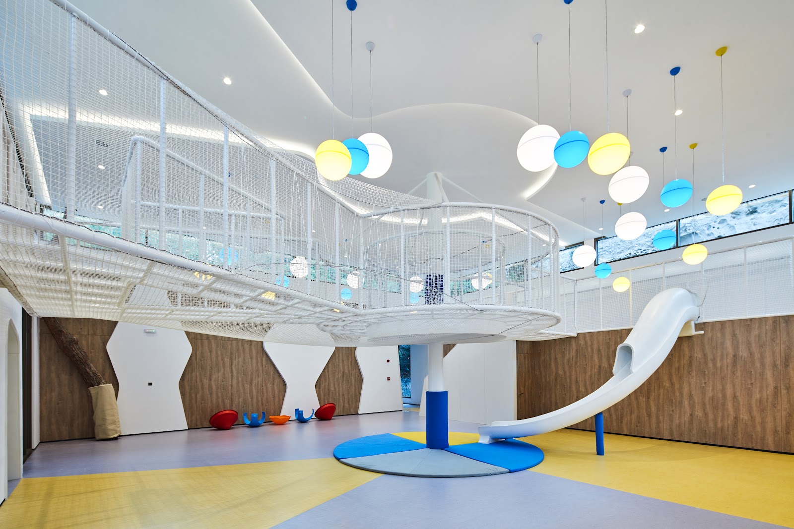 Modern lobby interior with geometric wooden ceiling panels, illuminated columns, patterned floor, and multi-story brick building visible through glass walls at night.