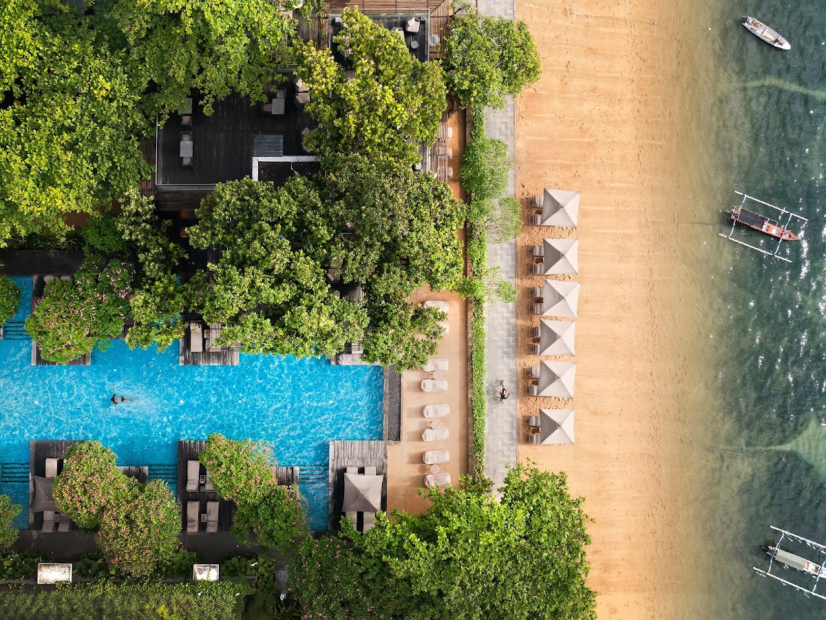 Aerial view of Maya Sanur Resort showing a beachfront pool, sandy beach with white umbrellas, lush green vegetation, and traditional boats in turquoise waters.