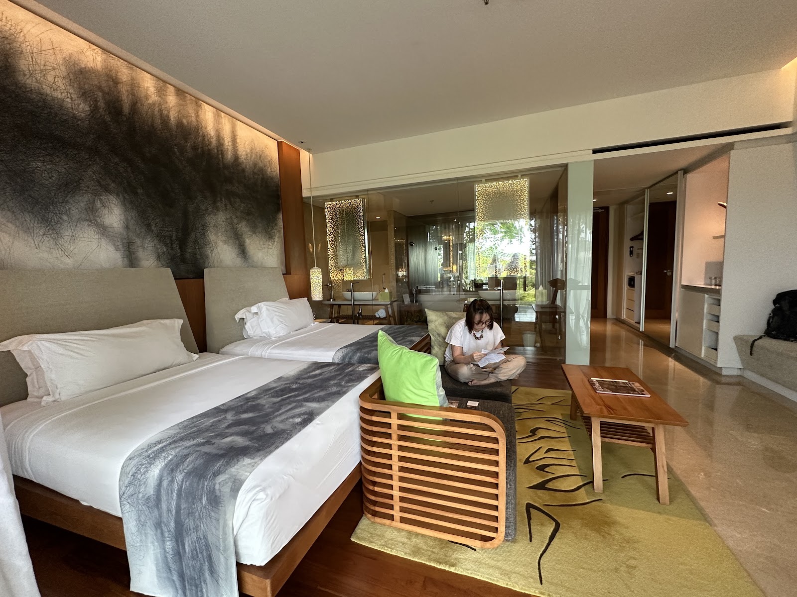 Modern resort suite with two beds, a guest seated in a green chair, and glass doors opening to a balcony overlooking tropical grounds.
