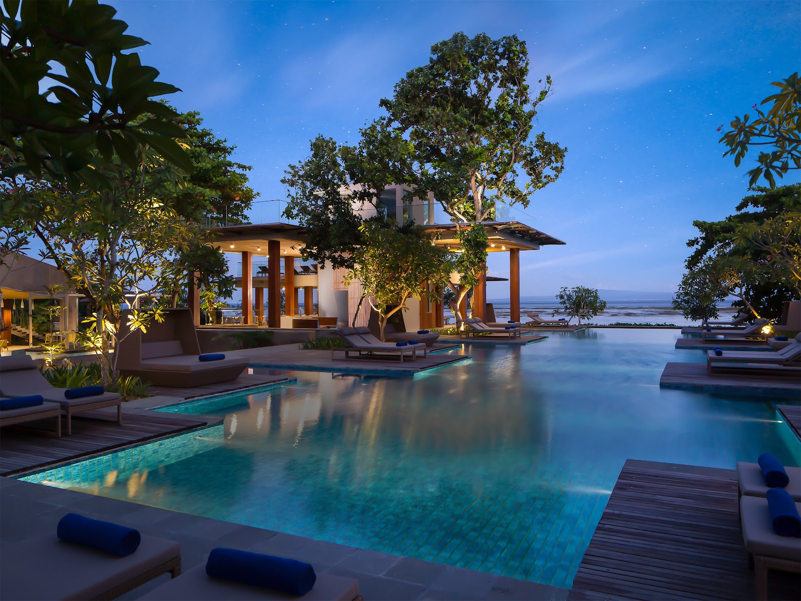 Illuminated pavilion with wooden pergola overlooks infinity pool at dusk, surrounded by lounge chairs and tropical trees at Maya Sanur Resort & Spa.