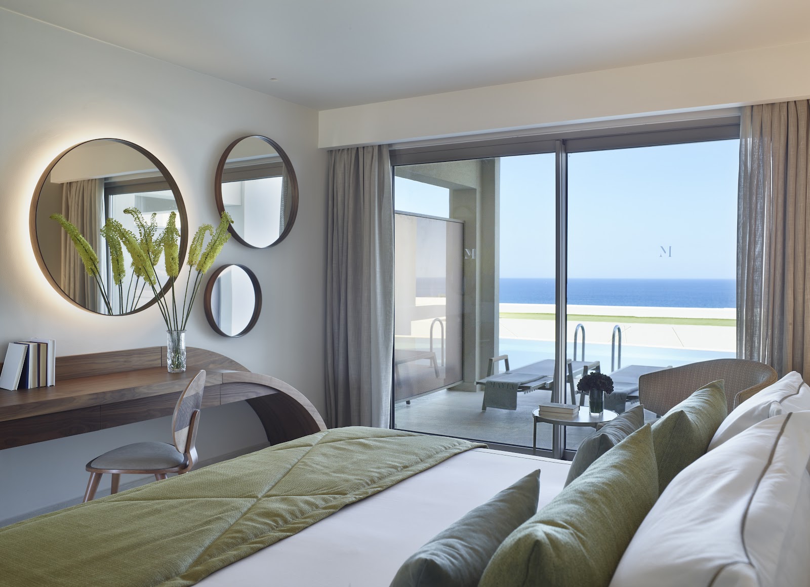 Resort suite bedroom with ocean view through sliding glass doors, bed with green bedding, round mirrors above desk, and pool visible beyond the terrace.