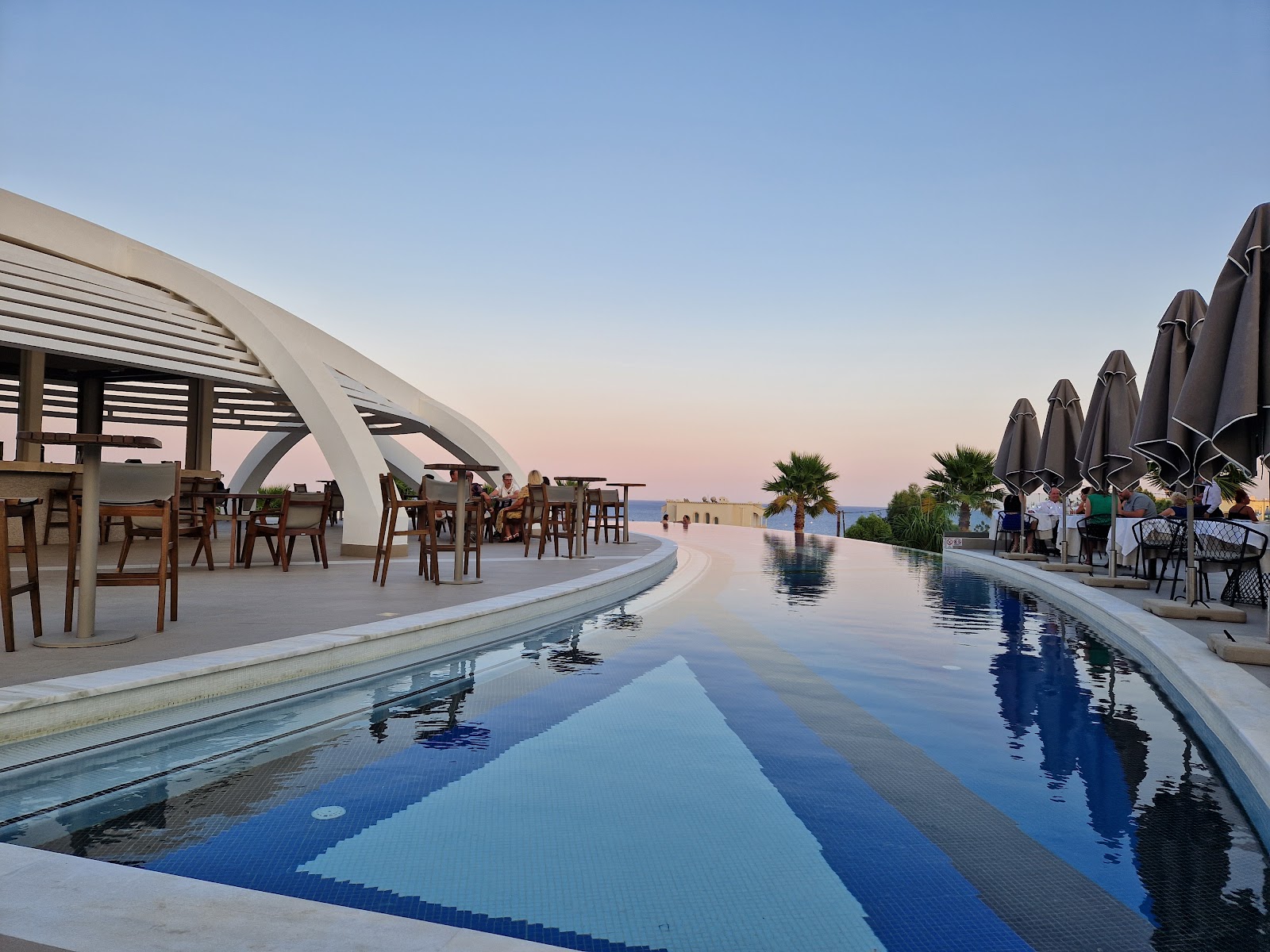 Infinity pool with geometric tile pattern at dusk, curved pergola and dining area on left, black umbrellas and guests on right, sea visible beyond.
