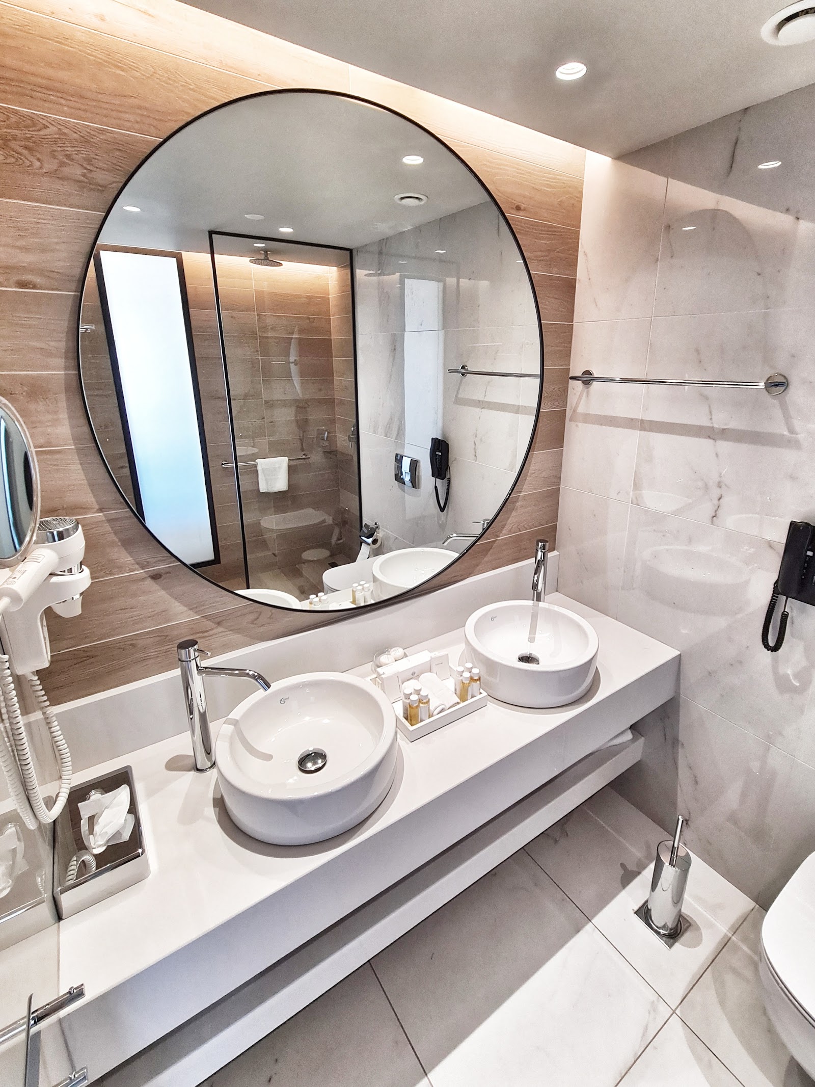 Modern bathroom with dual round white sinks, large circular mirror, wood and marble tile walls, and glass-enclosed shower.