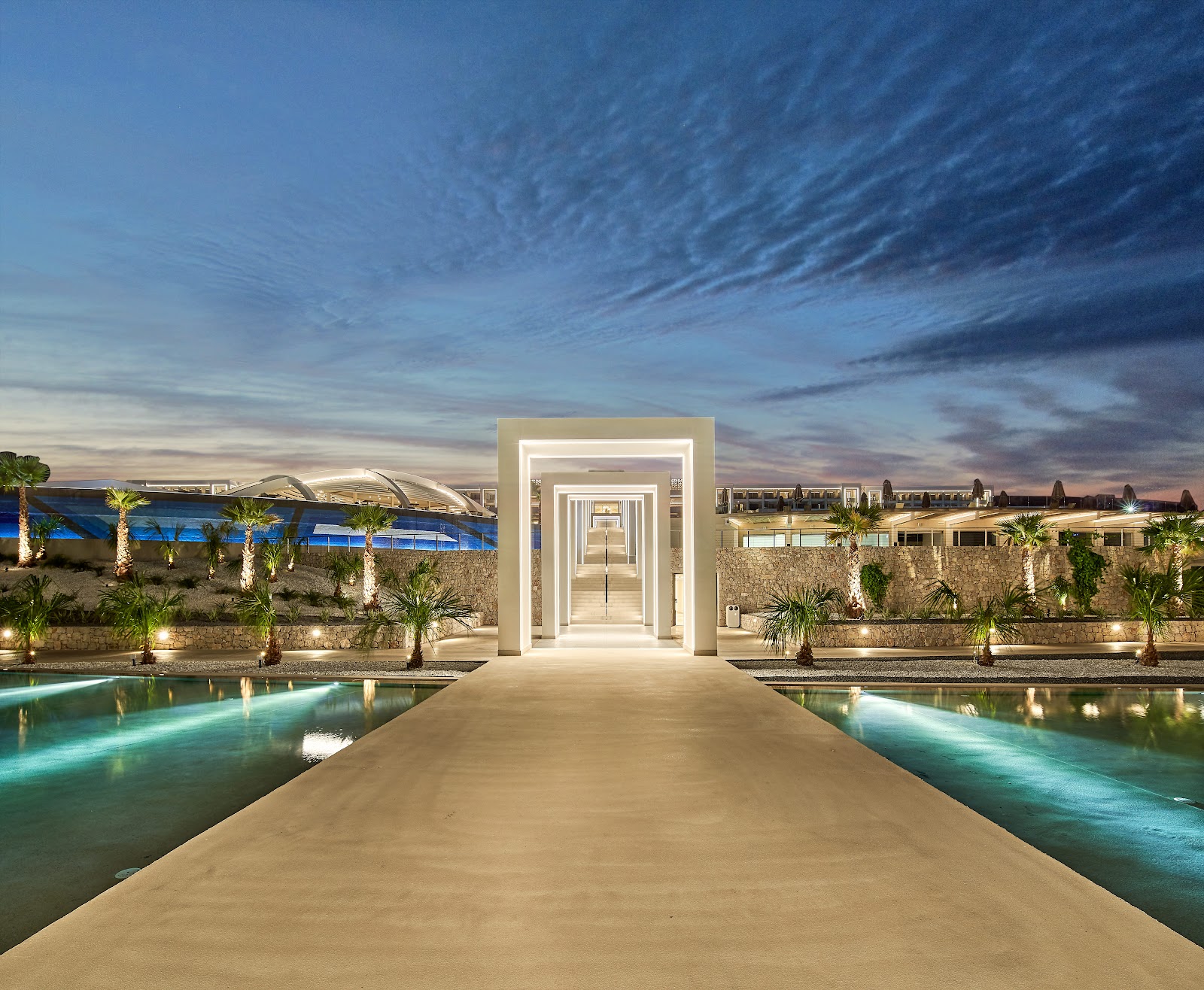 Mayia Exclusive Resort & Spa overlooks two large pools with palm trees, loungers, and a calm sea beyond under blue sky with white clouds.