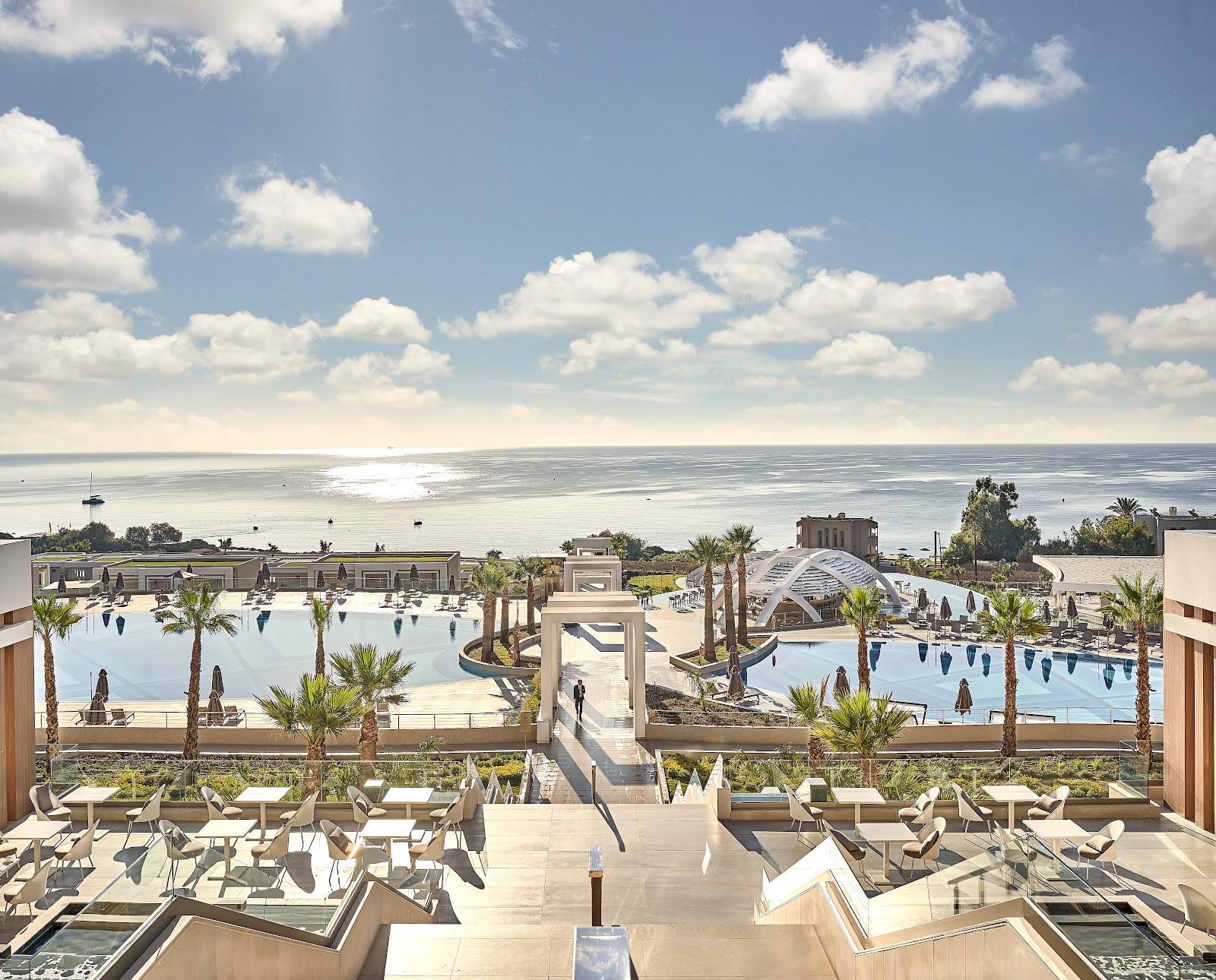 White modern resort building with geometric architecture framed by palm trees, lounge chairs visible on the beach terrace below.