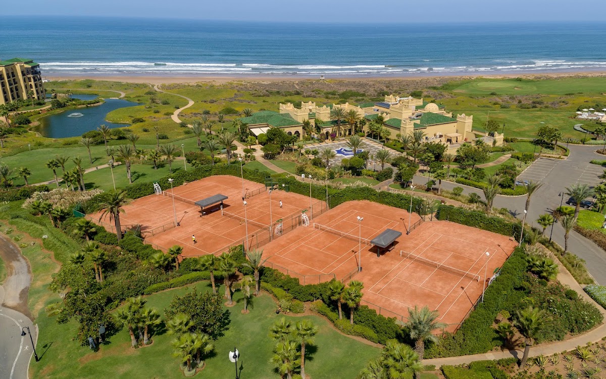 Clay tennis courts surrounded by palms and manicured grounds at Mazagan Beach & Golf Resort, with the ocean and beach visible beyond.