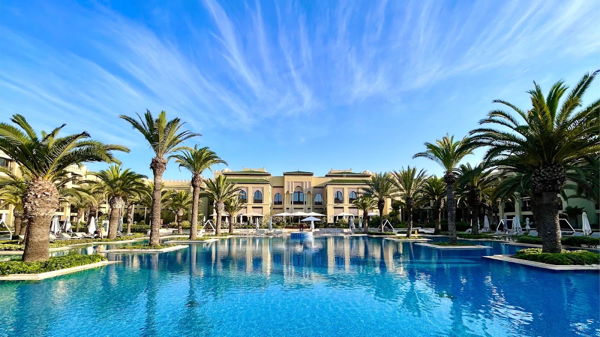 Large resort pool bordered by palm trees with tan buildings and white umbrellas under clear blue sky.