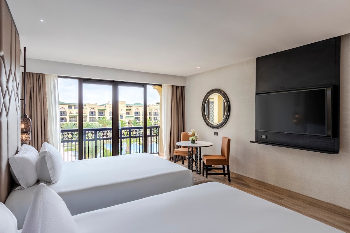 Resort guestroom with two queen beds, wall-mounted TV, and balcony overlooking lagoon and Moroccan architecture.