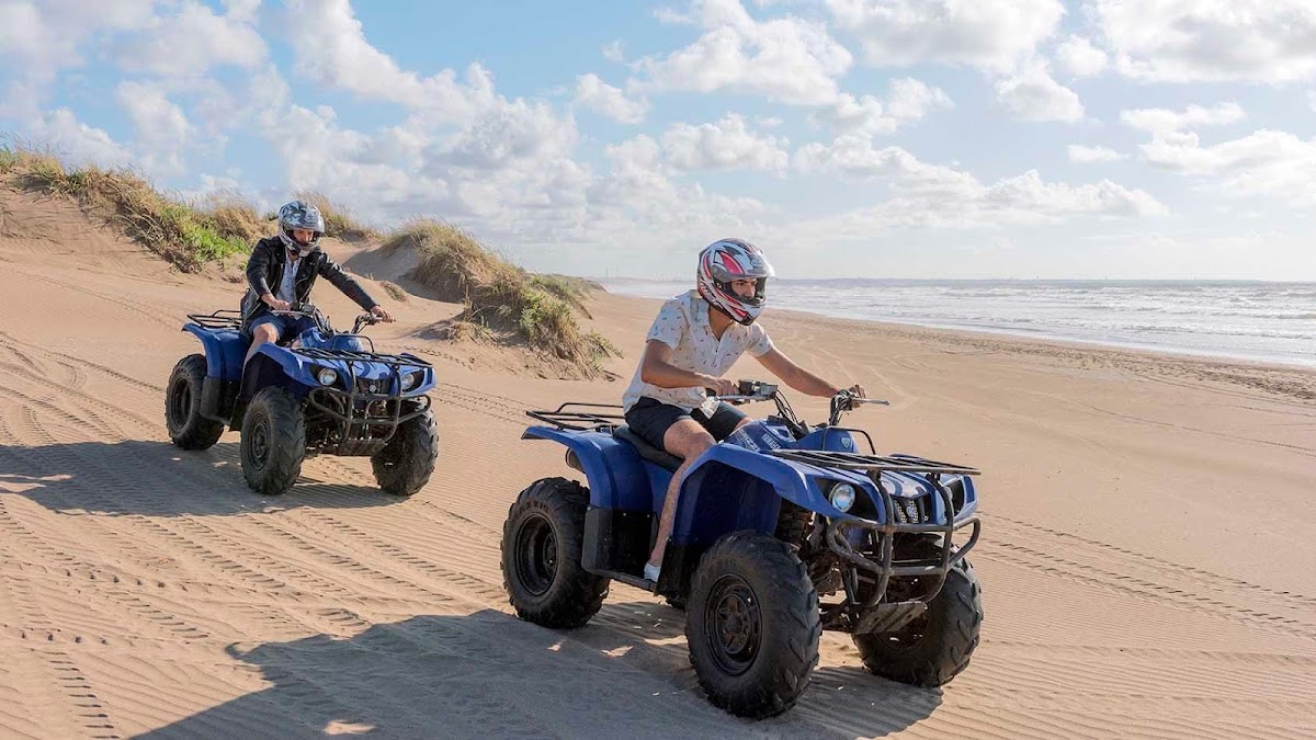 Two riders on blue all-terrain vehicles travel across a sandy beach with dunes and ocean in the background under a cloudy sky.