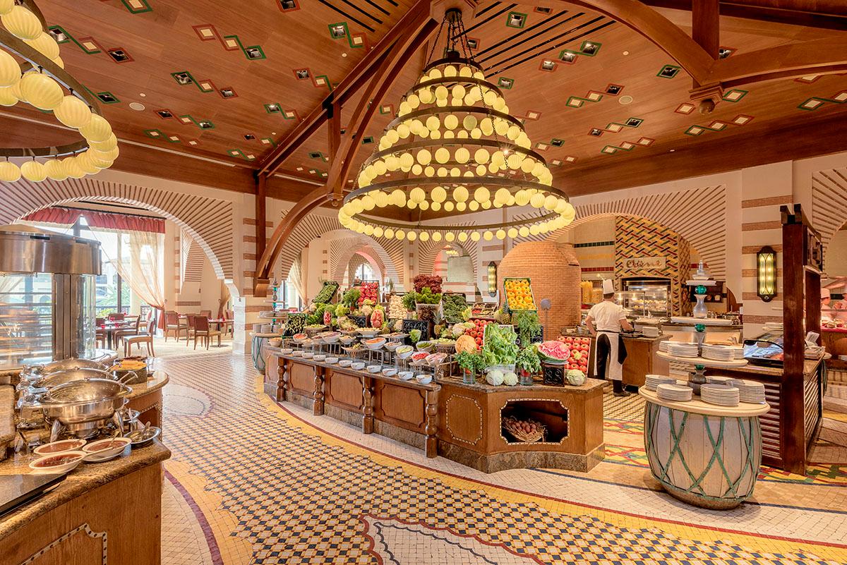 Mazagan Beach & Golf Resort's dining hall features a tiered chandelier, wooden beamed ceiling, and central food station decorated with fresh flowers and produce.