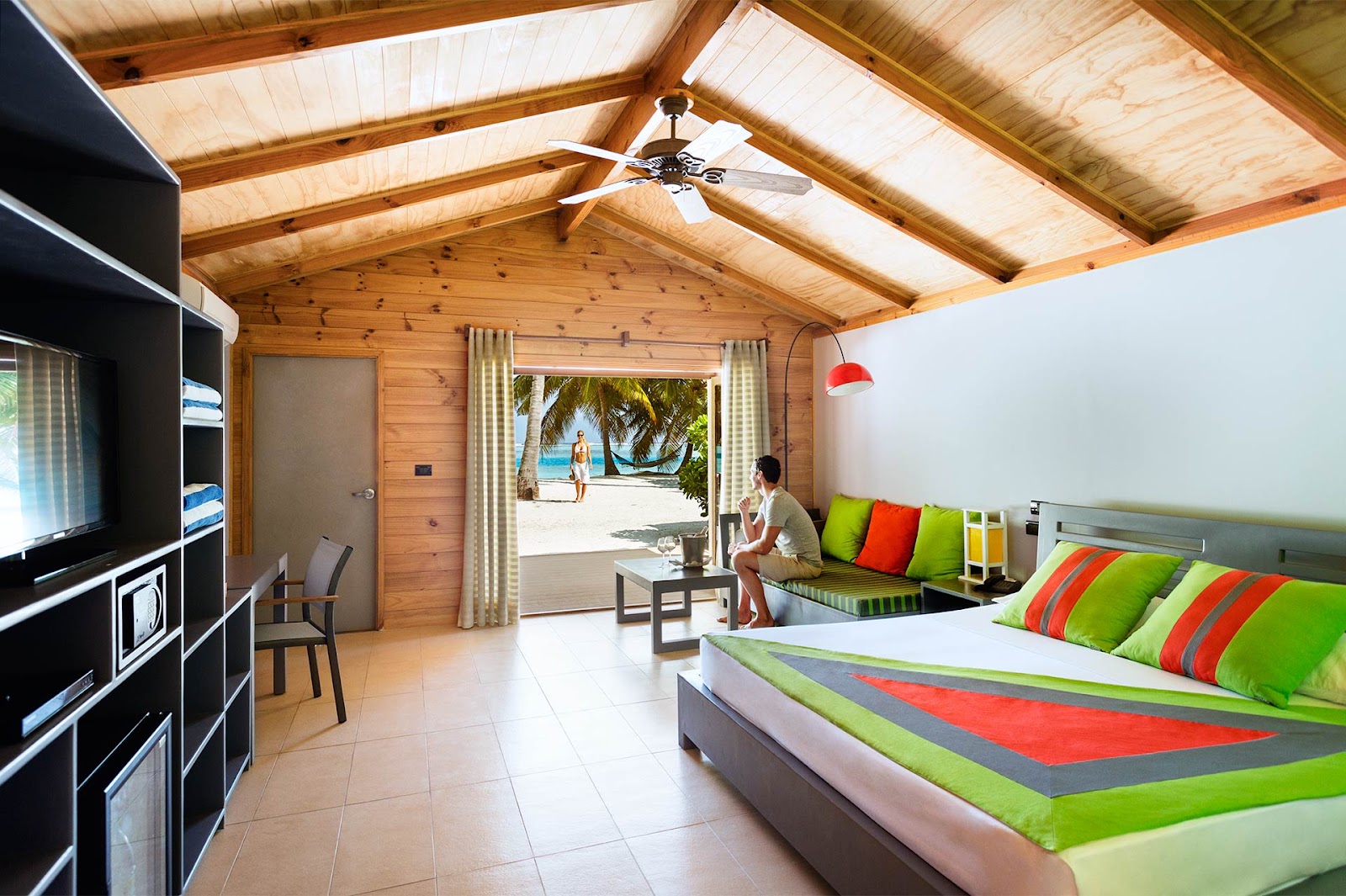Wood-beamed bungalow interior with bright green and orange bedding, open to a beach view with palm trees and ocean beyond.