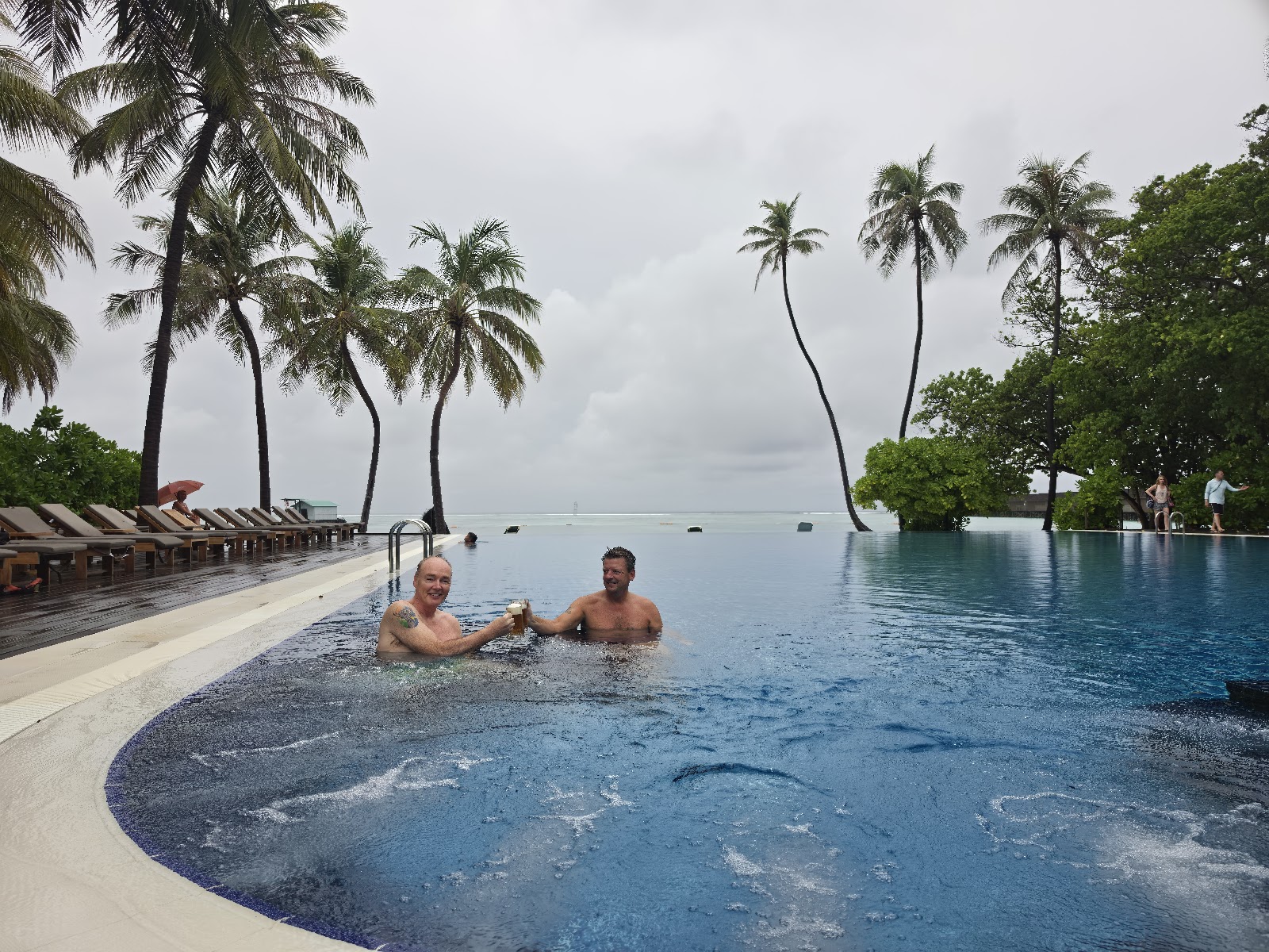 Two guests swim in an infinity pool overlooking the ocean at Meeru Maldives Resort, with palm trees and lounge chairs lining the deck.
