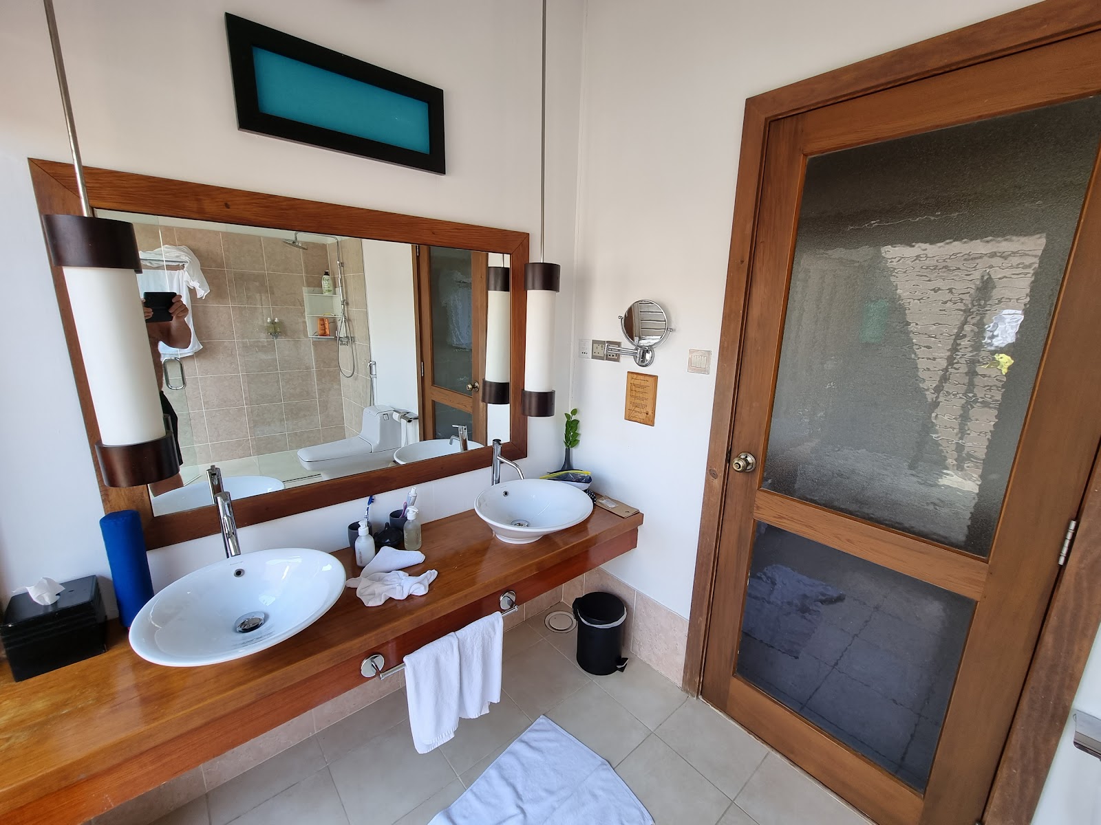 Resort bathroom with double sink vanity, wood-framed mirror, glass shower enclosure, and wooden door opening to ocean view.
