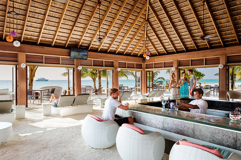 Resort pool with guests swimming, surrounded by palm trees and ocean view under blue sky.