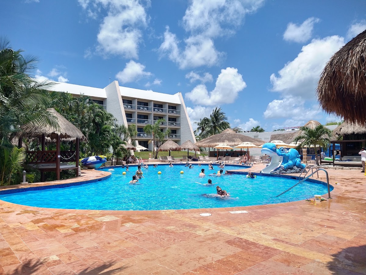 Guests swim in a bright blue pool with a blue elephant water slide, surrounded by thatched palapas and palm trees, with a modern hotel building in the background.