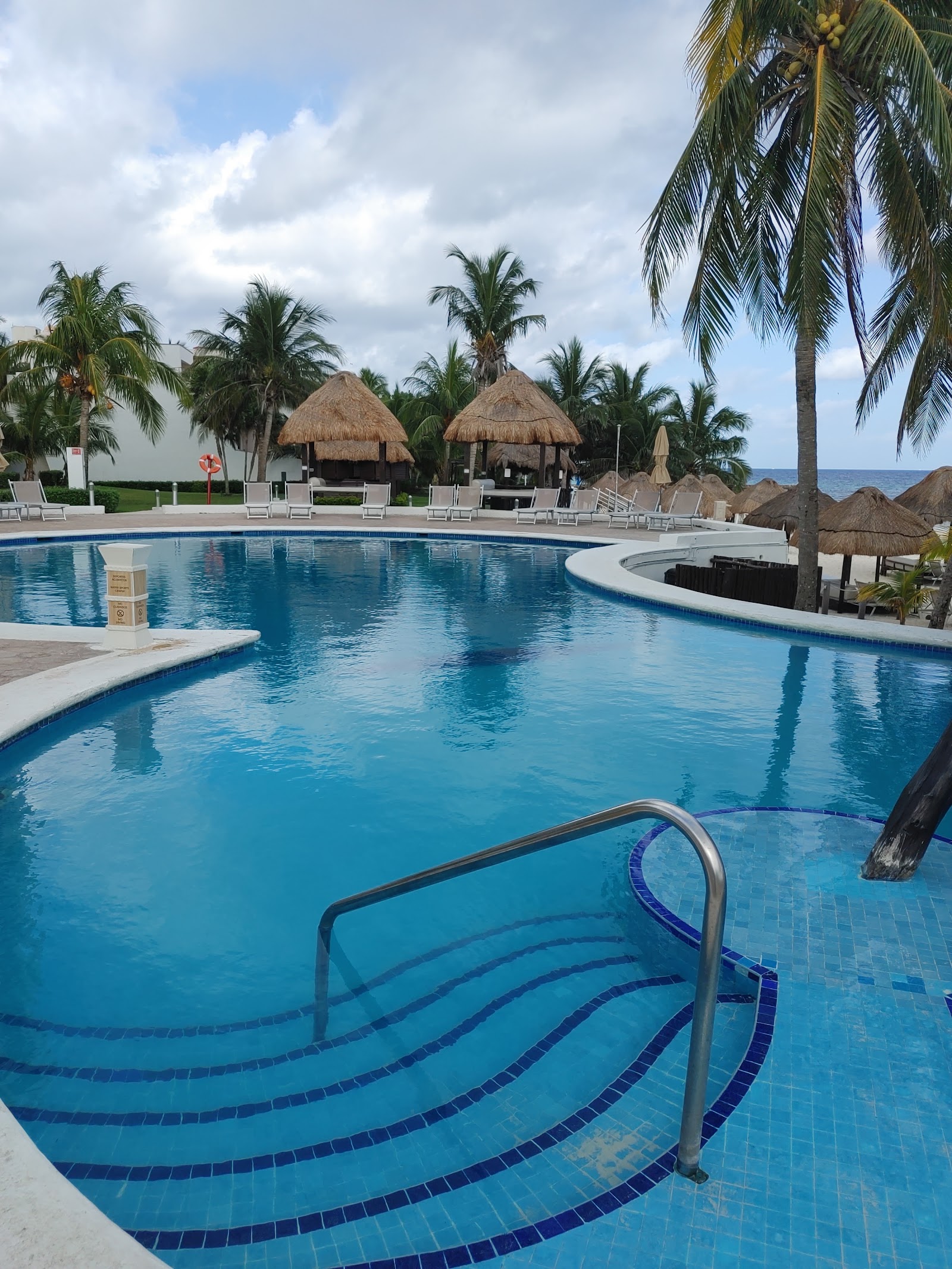 Resort pool with thatched-roof bar structures, palm trees, loungers, and ocean view beyond in the background.